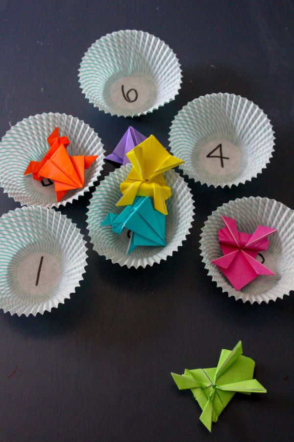 origami jumping frogs game in cupcake liners with numbers written in them and colored paper frogs, all on a black table