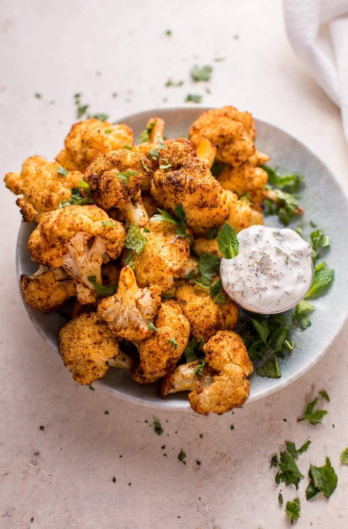roasted cauliflower in a white bowl with a small bowl of dip on a white table with some chopped greens on it