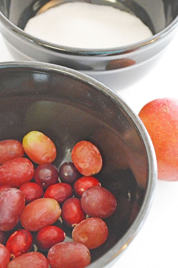 cranberries and grapes in a metal bowl next to another bowl of sugar next to a plum