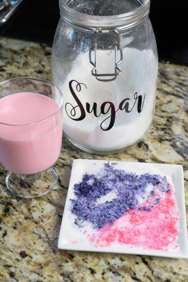 a glass of strawberry milk, a jar of sugar, a white plate of purple and pink colored sugar, on a kitchen counter