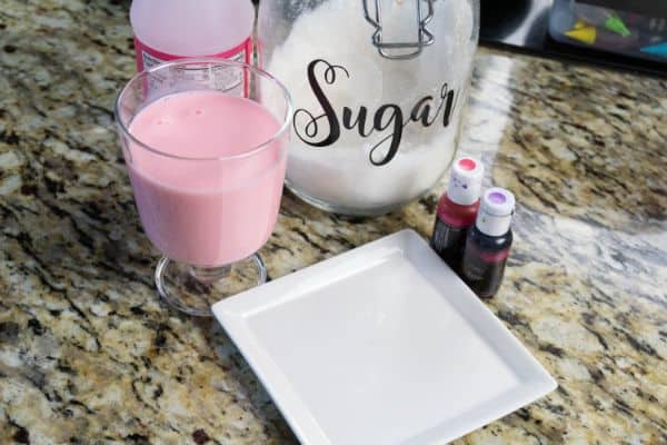 strawberry milk in a plastic container and in a glass, a glass jar of sugar, pink and purple dye, a white plate all on a kitchen counter