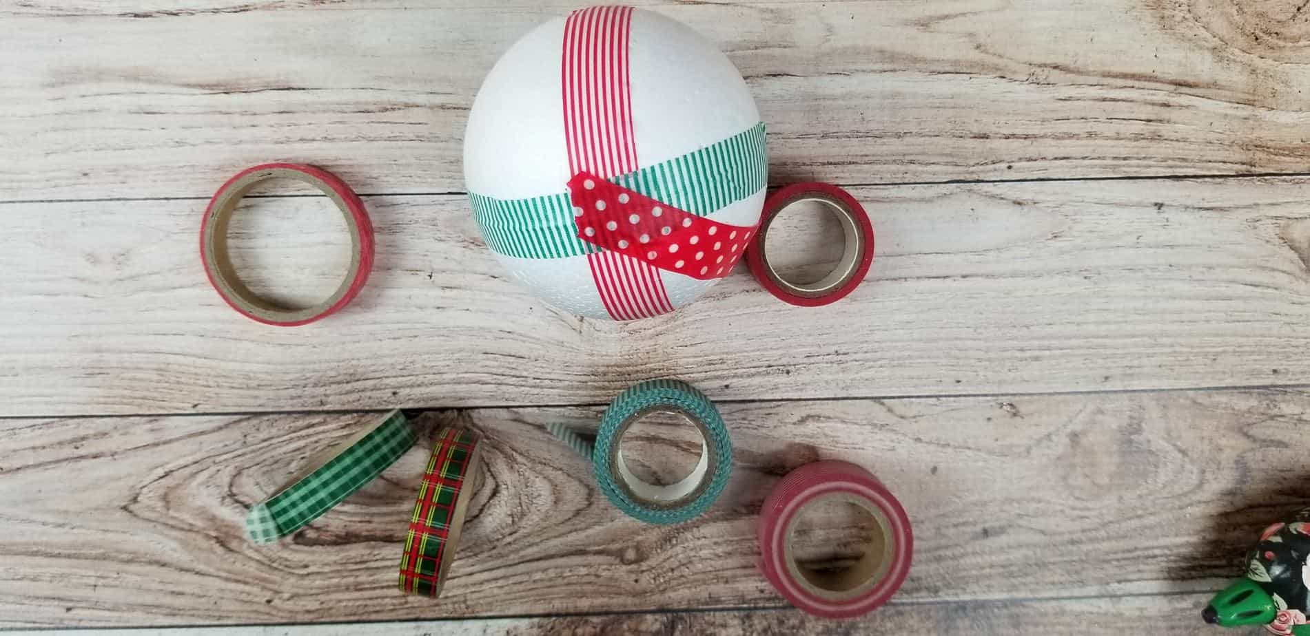 a styrofoam ball wrapped with some red and green washi tape with more washi tape rolls on the wooden table 