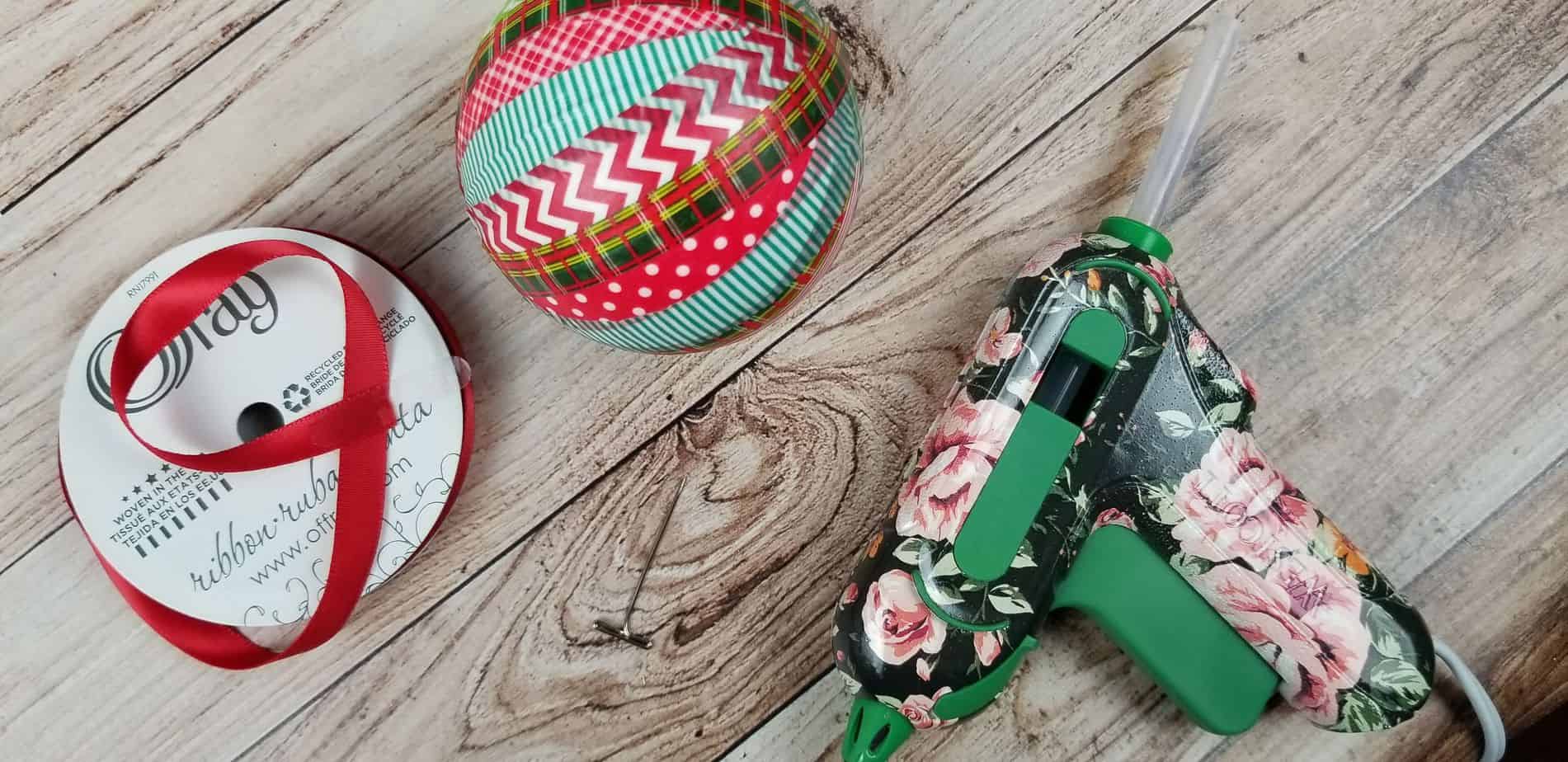 an ornament wrapped in red and green washi tape next to some red ribbon and a hot glue gun on a wooden table