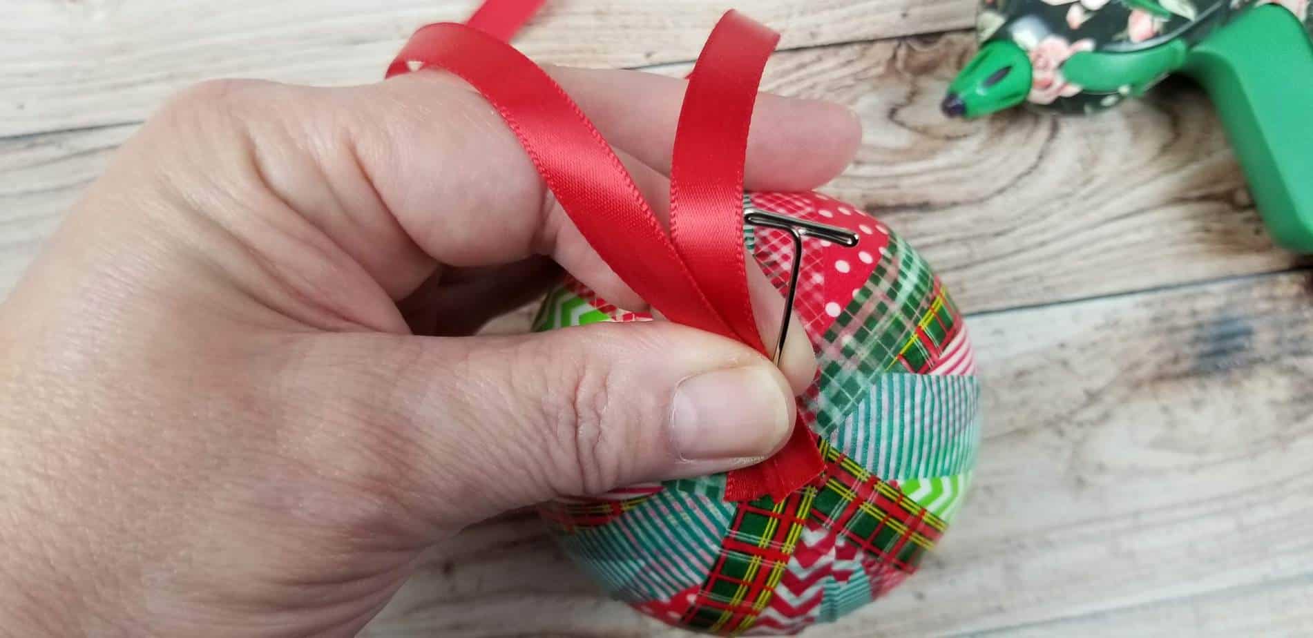 a hand holding a T pin and red ribbon at the top of the ornament wrapped in washi tape on a wooden table with a hot glue gun in the background