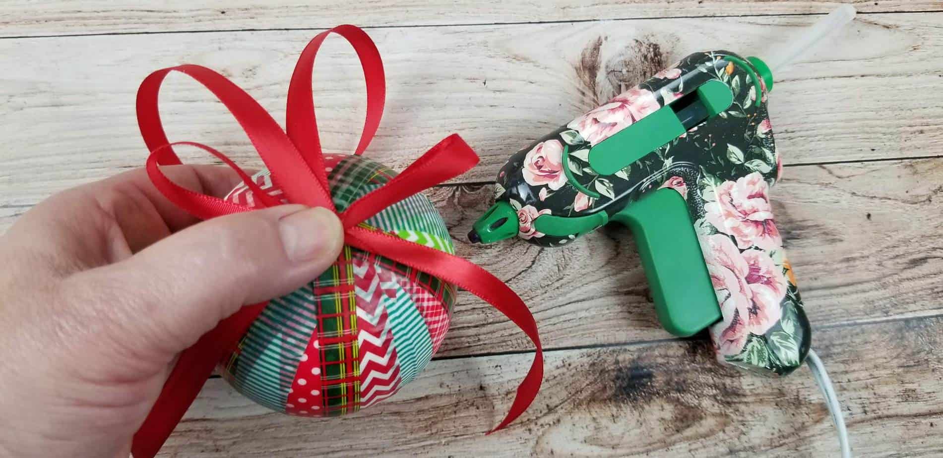 a hand holding red ribbon at the top of an ornament wrapped in washi tape on a wooden table next to a glue gun