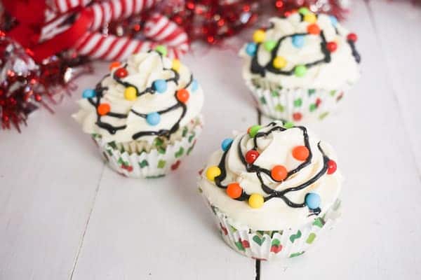 3 cupcakes topped with white frosting, multi-colored candy balls with black frosting in between to look like Christmas lights on a white table with candy canes in the background