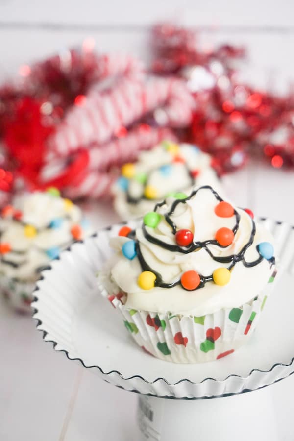 cupcake topped with white frosting, multi-colored candy balls with black frosting in between to look like Christmas lights on a cake plate with another cupcake and candy canes in the background