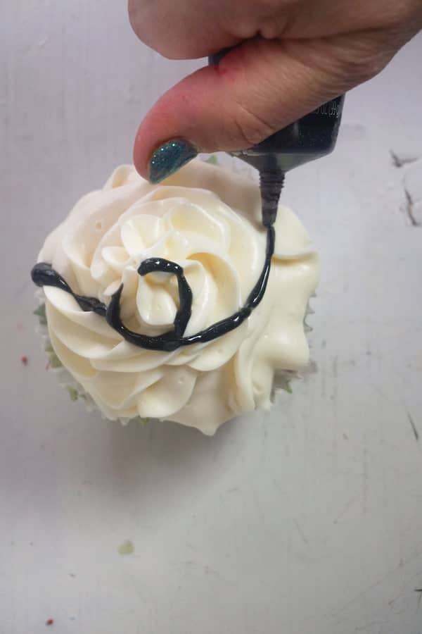 a hand swirling black writing gel on a cupcake topped with white frosting