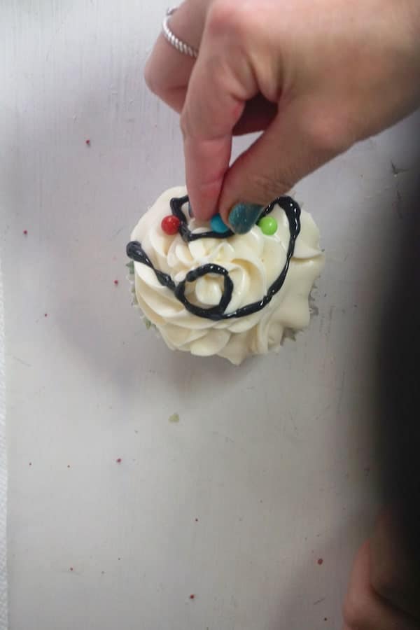 a hand placing colored round sprinkles on a cupcake topped with white frosting and black gel