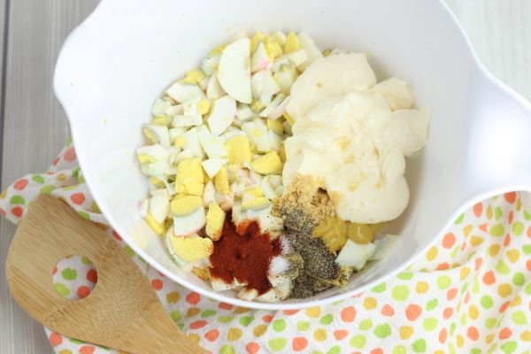 diced hard boiled eggs, mayonnaise, pickle juice, mustard, dry mustard, salt, pepper, onion powder and paprika in a white bowl on a multi-colored linen next to a wooden spoon on a gray wood table