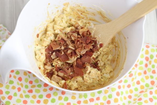 egg salad, diced bacon and a wooden spoon in a white bowl on a multi-colored linen on a gray wood table