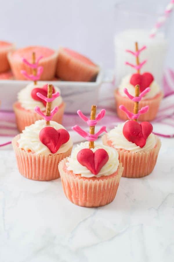 pink cupcakes topped with white frosting, a red heart, and a pretzel stick with pink frosting made to look like an arrow, all on a white counter with more cupcakes and a glass of milk in the background