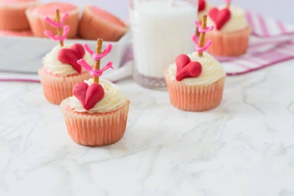 pink cupcakes topped with white frosting, a red heart, and a pretzel stick with pink frosting made to look like an arrow, all on a white counter with more cupcakes and a glass of milk in the background