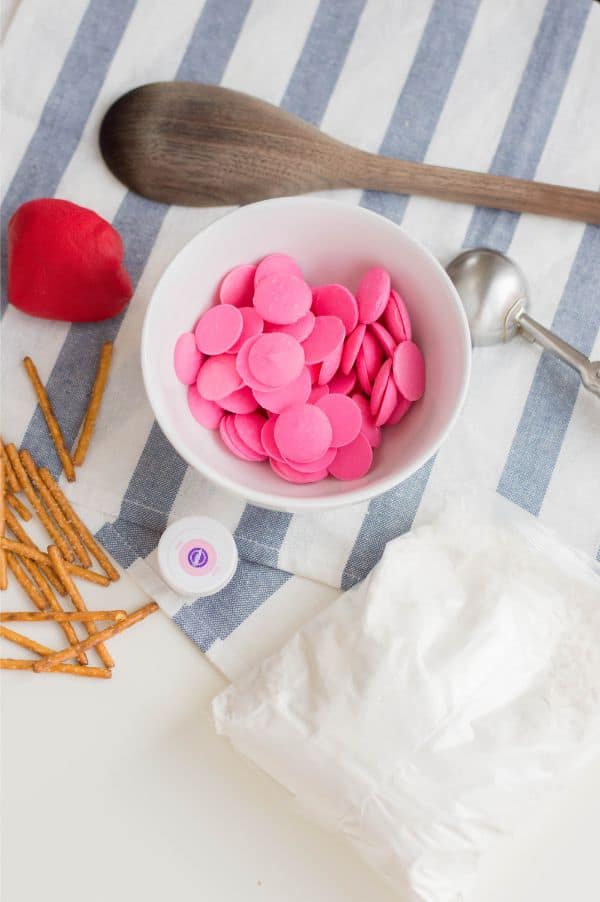 wooden spoon, red fondant, white bowl with pink candy melts in it, pretzel sticks, pink gel food coloring and confectioners sugar on a blue and white striped cloth