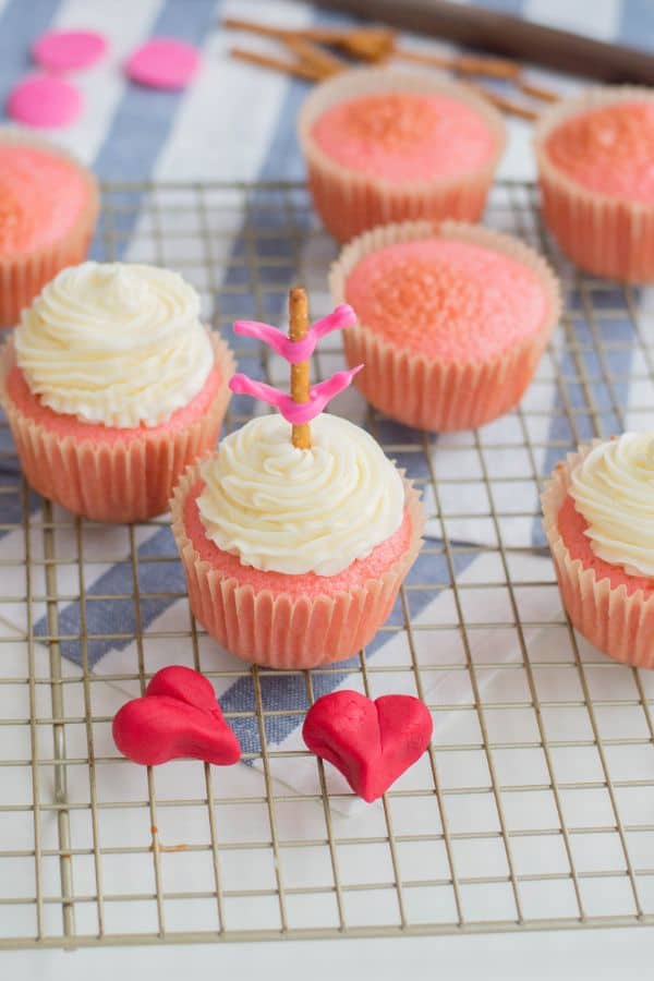 pink cupcakes, 3 topped with white frosting, and one topped with a pretzel stick with pink frosting on it to look like an arrow, next to two red fondant hearts on a wire rack on a blue and white striped cloth on a white table