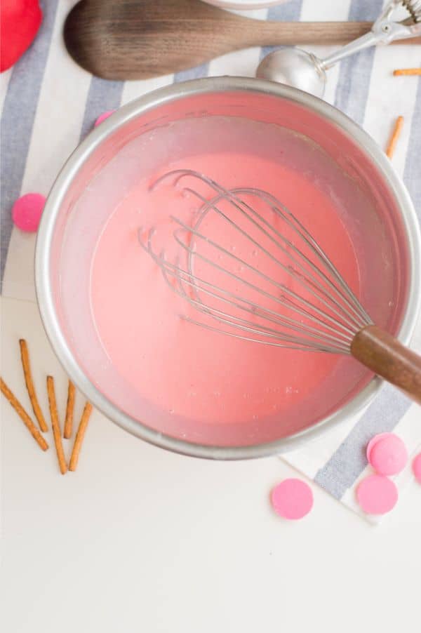 pink cake batter in a metal mixing bowl with a whisk in it next to a wooden spoon, pretzel sticks and pink candy melts on a blue and white striped cloth on a white table