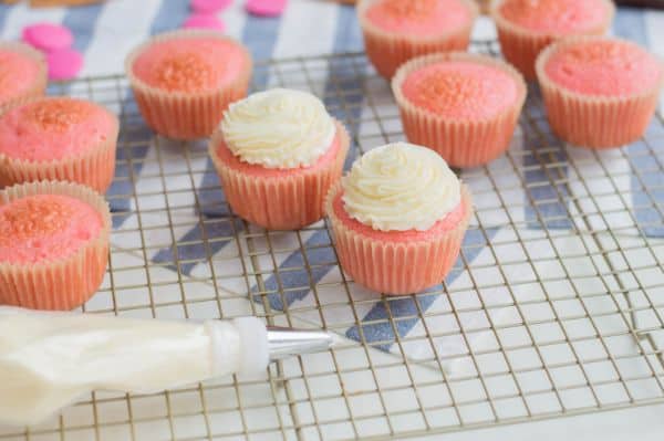 pink cupcakes, a couple with white frosting, the rest without, on a wire rack on a blue and white striped cloth on a white table