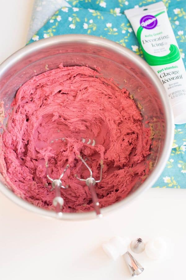 red frosting in a metal mixing bowl with metal mixers in it next to green decorating icing  and decorating tips on a blue and white flowered linen on a white counter