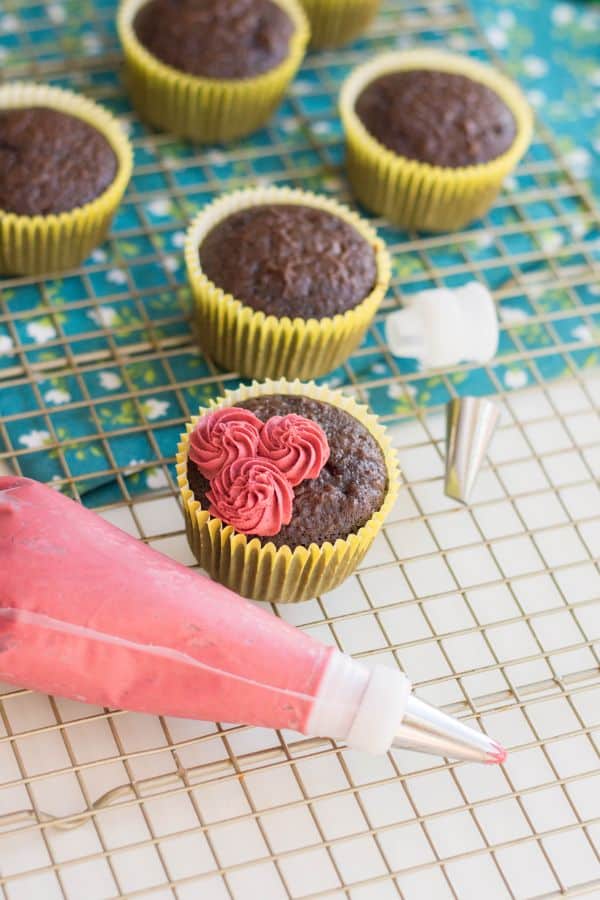 chocolate cupcakes, one starting to be topped with red frosting to look like roses, next to a bag of red frosting on a wire rack on a blue and white flowered linen on a white counter