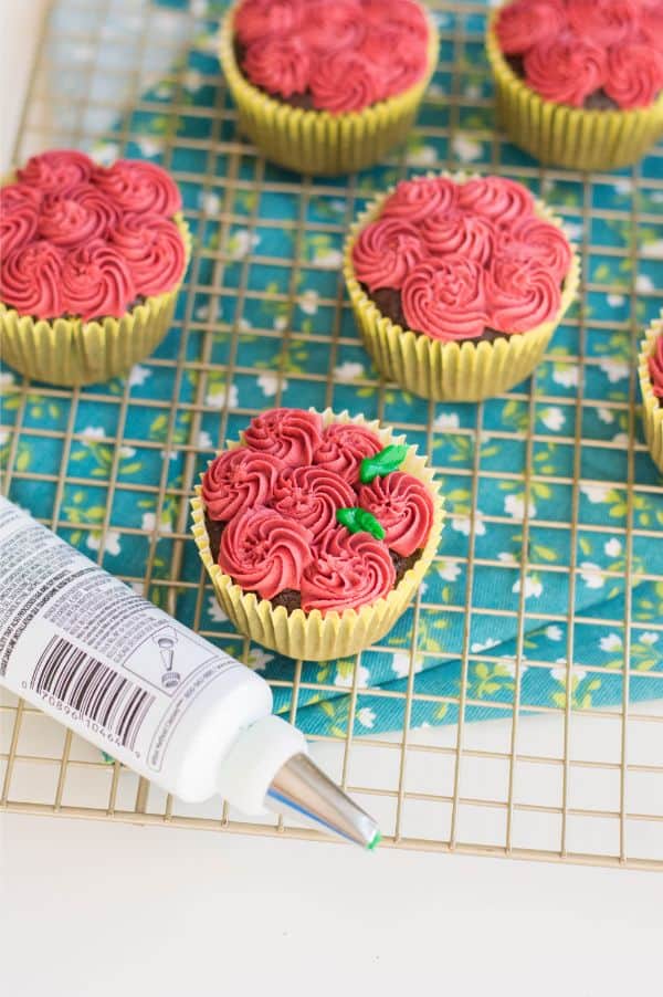 chocolate cupcakes decorated with red frosting to look like roses and green frosting for the stems on a wire rack next to a tube of frosting with a blue linen with white flowers underneath them