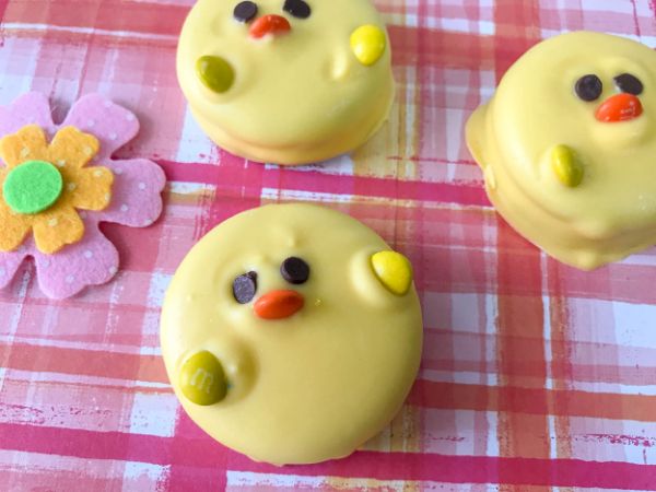 three oreos covered with yellow candy melts decorated with mini chocolate chips for eyes and M&Ms for the beak and wings so they look like Easter Oreo Chicks on a pink and orange cloth next to a felt flower