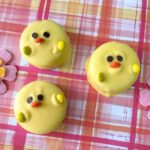 Three round yellow Easter Oreo Chicks, complete with orange beaks and black eyes, are nestled on a plaid surface adorned with pink floral accents.