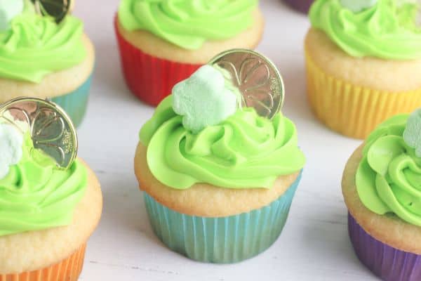 white cupcakes decorated with green frosting, a shamrock marshmallow and gold coin, on a wood table 