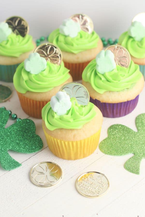 white cupcakes decorated with green frosting, a shamrock marshmallow and gold coin, on a wood table with more shamrocks, gold coins, and green beads on it
