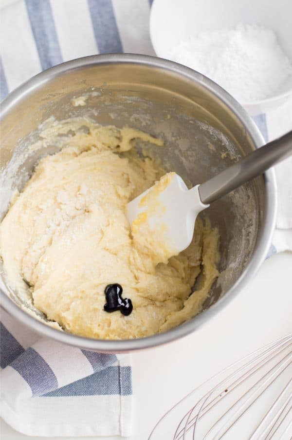 cake mix with drops of green food coloring on top of it in a metal bowl with a spatula in it next to a white bowl of powdered sugar on a white table next to a blue and white striped linen