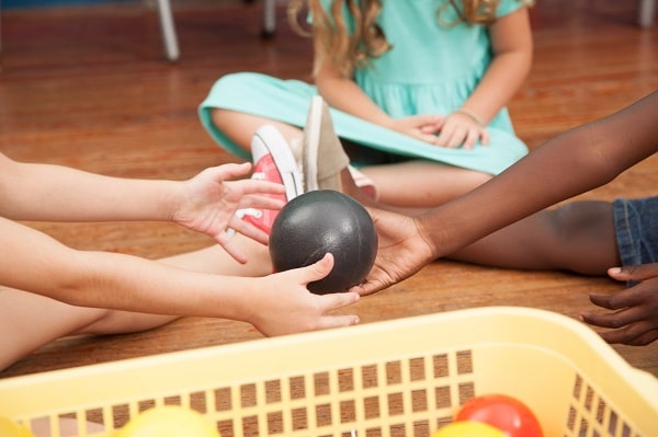 children's hands passing a black ball to each other with another girl sitting in the background on a wood floor, with a yellow basket with more balls in it next to the kids