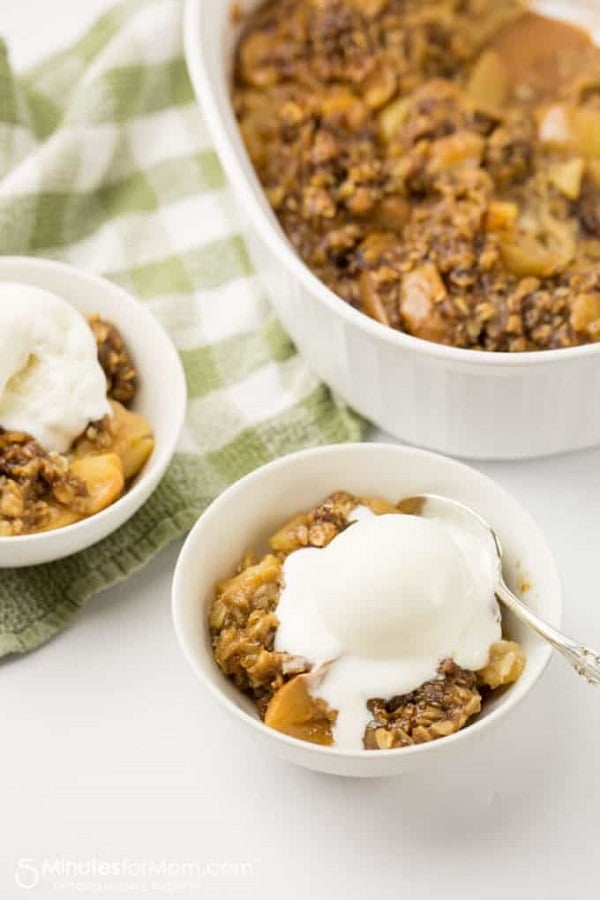 two white bowls of apple crisp topped with vanilla ice cream with a baking dish of apple crisp in the background on a white and green checkered linen