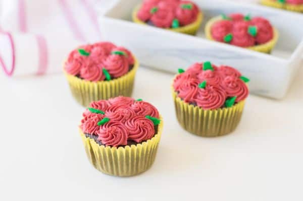 chocolate cupcakes decorated with red frosting to look like roses and green frosting for the stems on a white table with more cupcakes and a pink and white cloth in the background