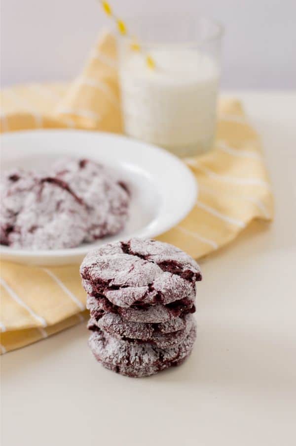 red velvet cookies stacked on a white table with more cookies on a white plate in the background on a yellow cloth next to a glass of milk