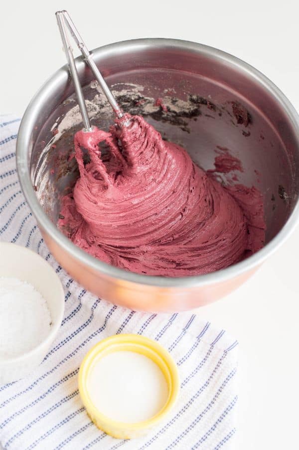 chocolate cake mix mixed with red gel food coloring in a metal bowl with beaters in it, next to a bowl of granulated sugar and confectioners sugar on a blue and white cloth on a white table