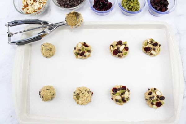 trail mix cookie dough balls on baking sheet next to a cookie scoop and glass bowls of more trail mix ingredients