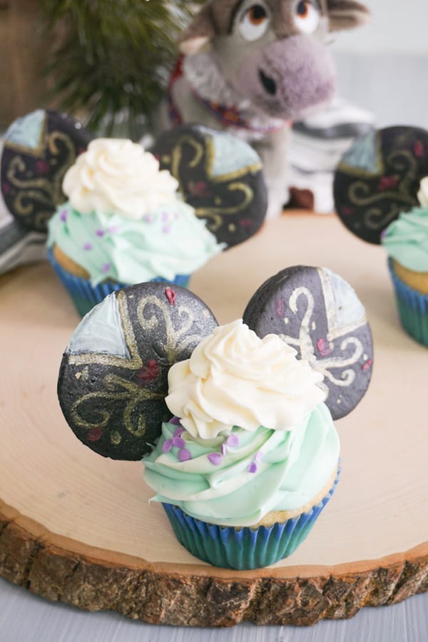 closeup of cupcakes decorated with green ans white frosting and decorated cookies to look like Princess Anna Mickey Ears cupcakes on a log