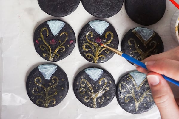 a hand painting white and gold icing with a paintbrush on  black fondant circles