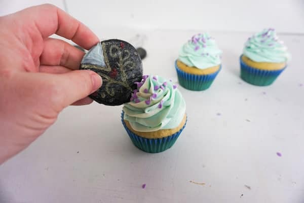 a hand putting a decorated black fondant circle on a cupcake decorated with blue frosting and purple sprinkles with two more decorated cupcakes in the background
