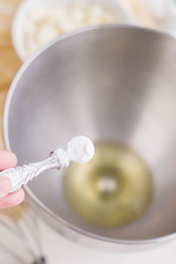 a hand holding the cream of tartar in a measuring spoon above a metal bowl with egg whites in it