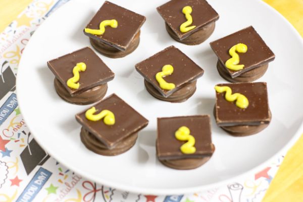 oreos covered in chocolate topped with a square chocolate candy and yellow frosting to look like Graduation Cap Cookies on a white plate on a graduation celebration paper
