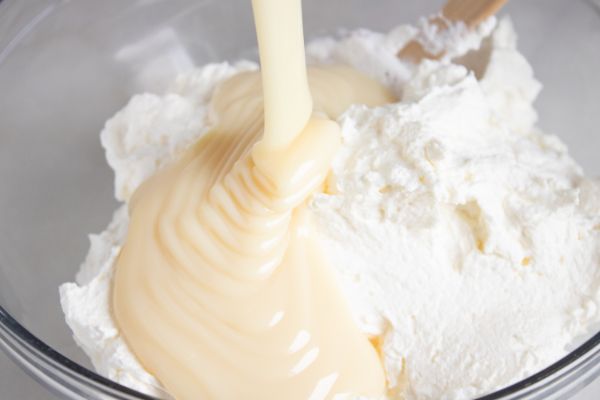 sweetened condensed milk being poured into a glass bowl of mixed heavy whipping cream