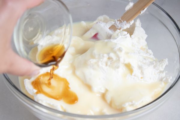 vanilla extract being poured into a glass bowl of sweetened condensed milk and mixed heavy whipping cream