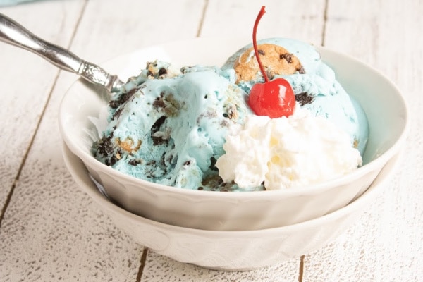 homemade cookie monster ice cream topped with a cherry in a white bowl with a spoon in it on a white table