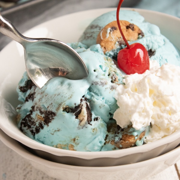 homemade cookie monster ice cream topped with a cherry in a white bowl with a spoon in it with more ice cream in a pan in the background