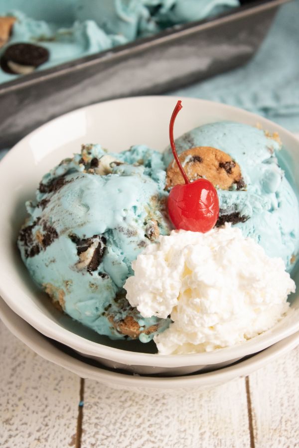 homemade cookie monster ice cream topped with a cherry in a white bowl with more ice cream in a pan in the background