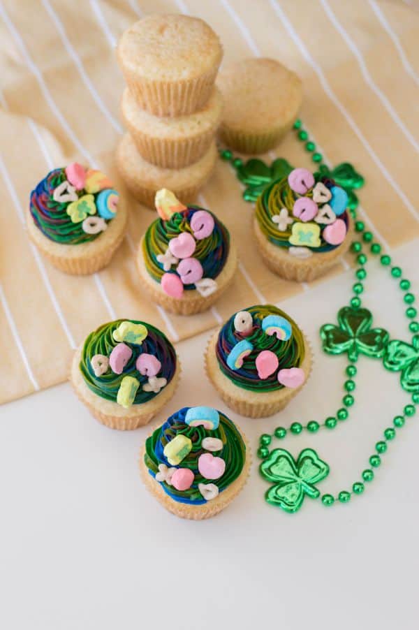 overhead view of vanilla cupcakes with multi-colored frosting topped with lucky charms marshmallows next to green shamrock beads on a tan linen with stacked vanilla cupcakes in the background