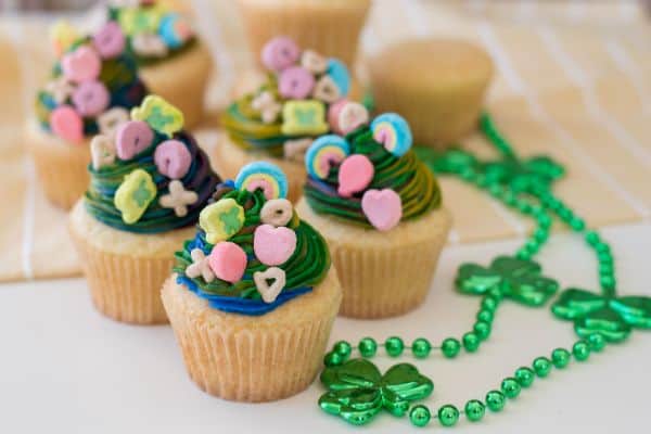 vanilla cupcakes with multi-colored frosting topped with lucky charms marshmallows next to green shamrock beads on a tan linen with stacked vanilla cupcakes in the background