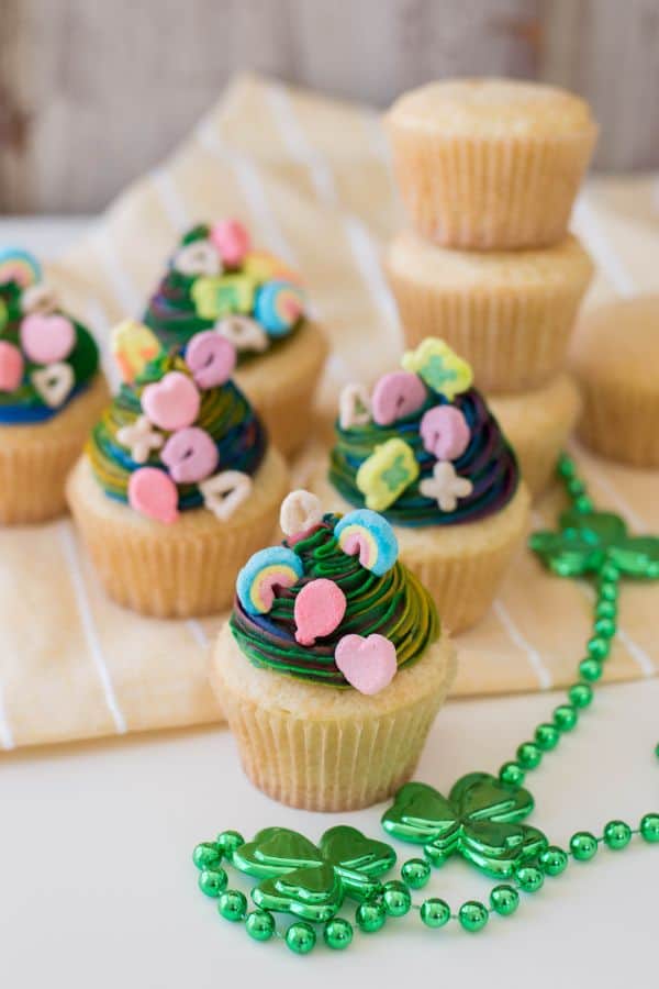 vanilla cupcakes with multi-colored frosting topped with lucky charms marshmallows next to green shamrock beads on a tan linen with stacked vanilla cupcakes in the background