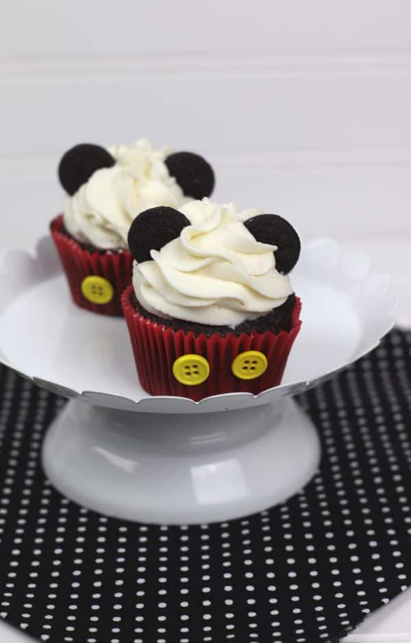 chocolate cupcakes decorated with white frosting, mini oreos, and two yellow buttons on the red cupcake liner, to look like Mickey Mouse, all on a white cake plate on a black and white polka dot cloth on a white background