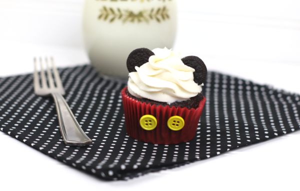 chocolate cupcake decorated with white frosting, mini oreos, and two yellow buttons on the red cupcake liner, to look like Mickey Mouse,  on a black and white polka dot cloth on a white background next to a fork and a glass of milk